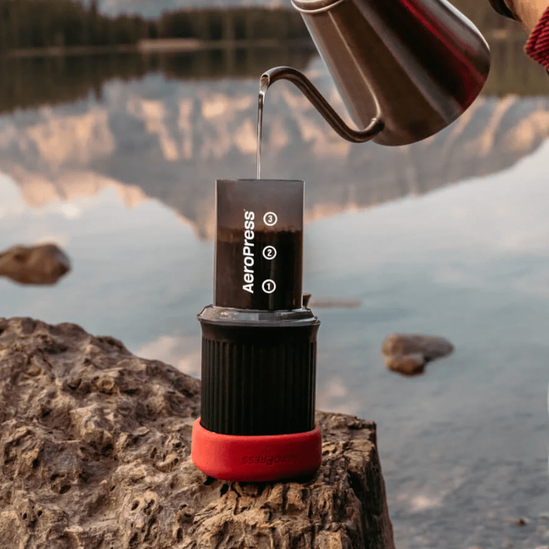 The AeroPressGo brewing coffee on a rock in front of a lake.